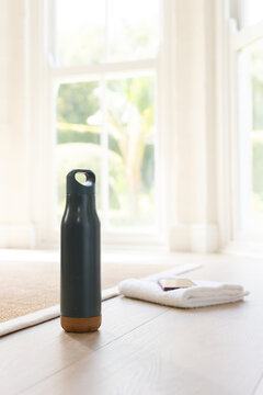 Dark green reusable bottle is standing on wood floor near tall windows, towel and phone nearby