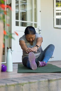 Senior female stretching reaching left leg on porch green mat in sportswear with water bottle