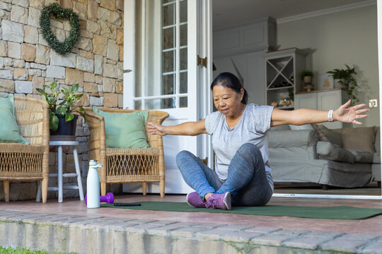 Senior Asian woman sitting on porch mat raising arms in gray tee near white bottle purple-dumbbell