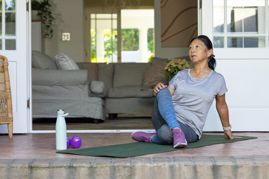 Senior Asian woman in grey-top performing seated twist on green mat on porch, dumbbells, copy space