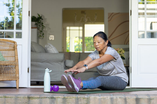 Senior Asian woman stretching forward in workout clothes on mat at home entry with purple dumbbells