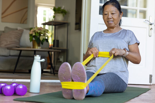Senior Asian woman performing seated yellow resistance band exercise on green mat in living room