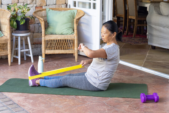 Senior Asian woman exercising on green mat pulling yellow band, wearing grey leggings, copy space