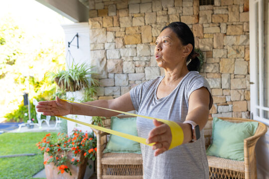 Senior Asian woman stretching yellow resistance band on porch by green-cushioned bench