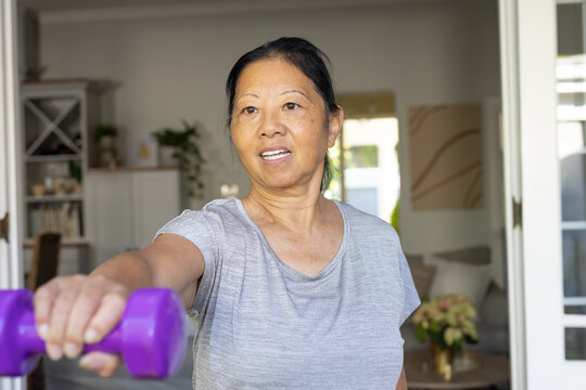 Senior Asian woman lifting purple dumbbell in home living room, wearing light gray T-shirt