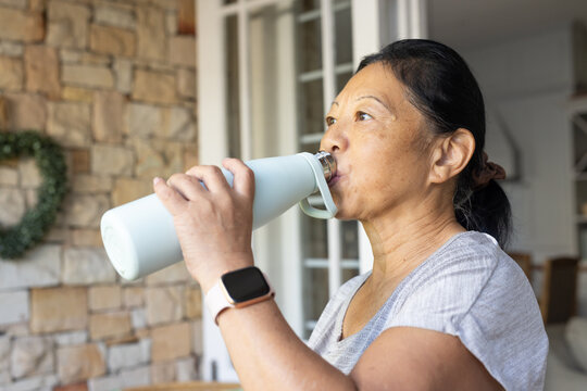 Senior Asian woman drinking from blue bottle wearing square smartwatch near French doors at home