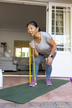Senior Asian woman exercising, pulling yellow band on green mat on tiled porch, wearing gray tee