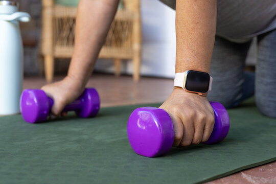 Senior woman kneeling on green mat gripping purple dumbbells at home wearing pale band smartwatch