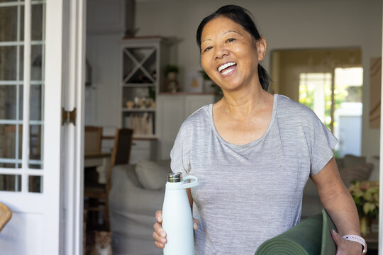 Senior Asian woman standing at home holding blue bottle, green yoga mat, wearing fitness tracker