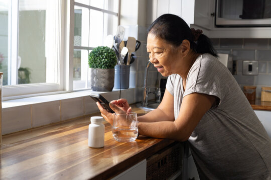 Senior Asian woman leaning in kitchen by window holding phone, pill bottle, glass, copy space