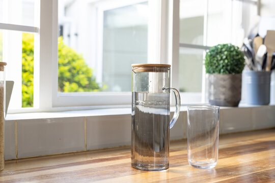 Tall clear glass carafe is sitting on wooden countertop near white window, with empty glass