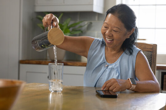 Senior Asian woman pouring water from pitcher into glass at kitchen table, phone, light-blue blouse