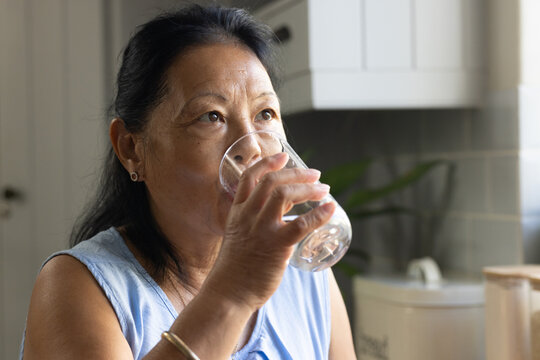 Senior Asian woman standing at kitchen counter in light blue top, sipping water from clear glass