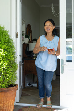 Senior Asian woman standing in front doorway holding clear glass cup of tea, near terracotta pot