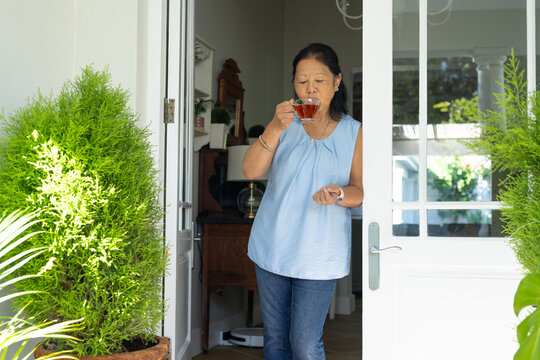Senior Asian woman standing sipping tea in clear glass teacup at entryway in blue blouse, jeans
