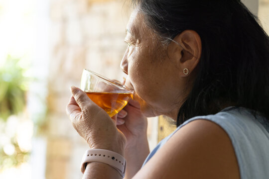 Senior Asian woman holding clear glass teacup and wearing wristband while sipping tea near window