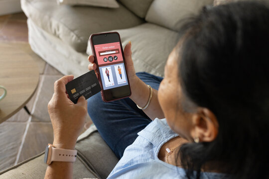 Senior Asian woman holding red-case smartphone and black chip card, shopping on beige sofa