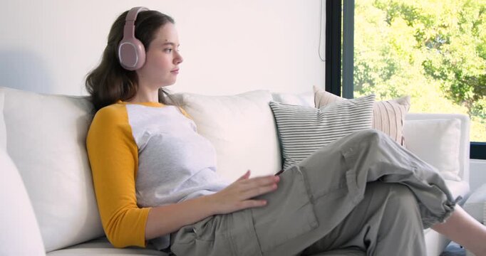 Adult woman resting on white sofa by large window, wearing pink headphones and using red smartphone