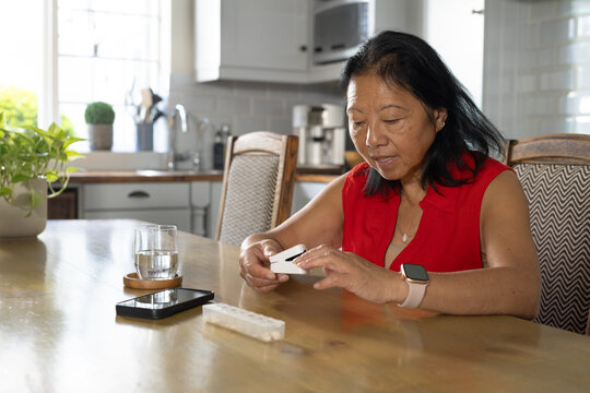 Senior Asian woman in red blouse examining device at kitchen table with pill tray, copy space