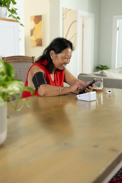 Senior Asian woman wearing red sleeveless top using tablet at dining table with blood pressure cuff