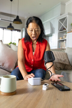 Senior Asian woman sitting at home in red sleeveless top, blue jeans checking cuff, BP monitor