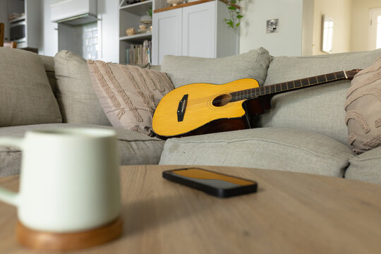 Yellow acoustic guitar is resting diagonally on grey sofa near coffee table, mug, smartphone, plant