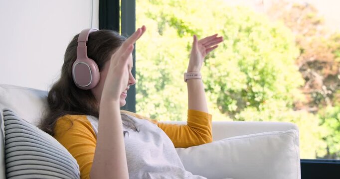 Female with pink headphones listening on white sofa raising hands reaching for smartphone scrolling