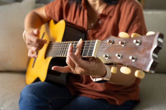 Senior woman playing yellow-top acoustic guitar on sofa at home, wearing rust polo, wristwatch