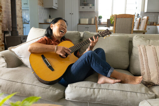 Senior Asian woman sitting on light-gray sofa playing acoustic guitar and singing at home