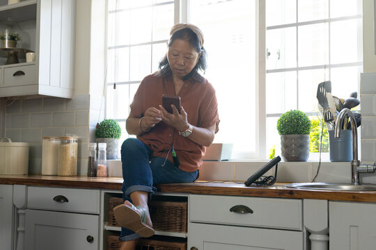 Senior Asian woman sitting on kitchen counter holding smartphone with earbud cord and tablet