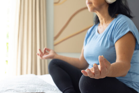 Senior Asian female sitting cross-legged on bed meditating with white headphones and black leggings