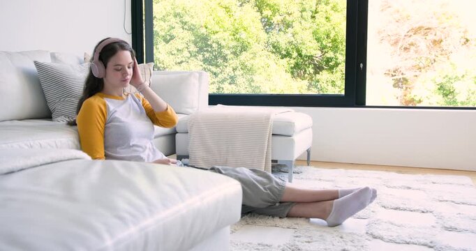 Woman sitting in living room leaning on sofa, adjusting pink headphones as music starting on device