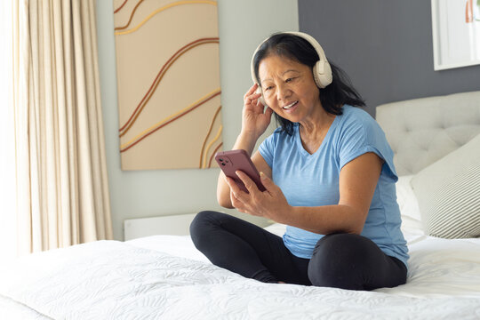 Senior Asian woman sitting on bed wearing white headphones and holding pink-cased smartphone