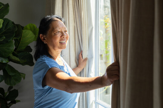 Senior woman in blue-shirt pulling beige curtains at home by window with plant letting sunlight in