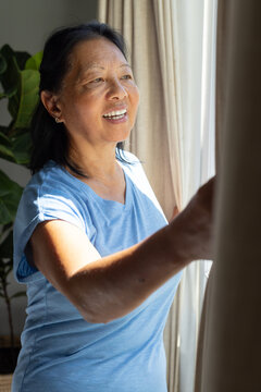 Senior Asian woman standing by sunlit window at home pulling beige curtains, showing stud earrings