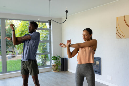African American couple in sportswear stretching arms across chest in living room by rolled mats