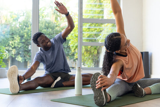 African friends in sportswear doing seated side stretches on green mats at home with water bottle