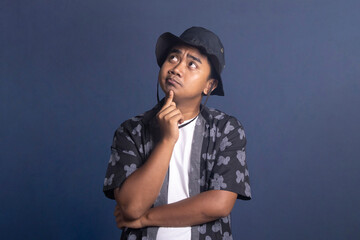 Young man wearing a black bucket hat and patterned shirt looking upward with a thoughtful and pensive expression while touching his chin. © Reza
