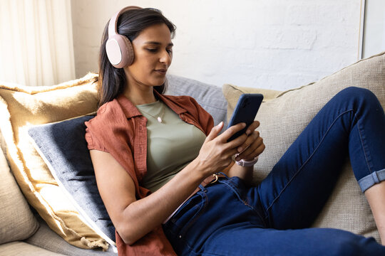 Adult Indian woman reclining on beige sofa in living room, using smartphone and wearing headphones
