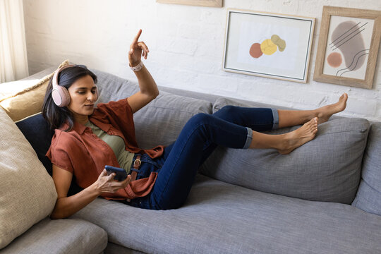 Indian woman reclining at home on gray sofa, listening with pink headphones, holding smartphone