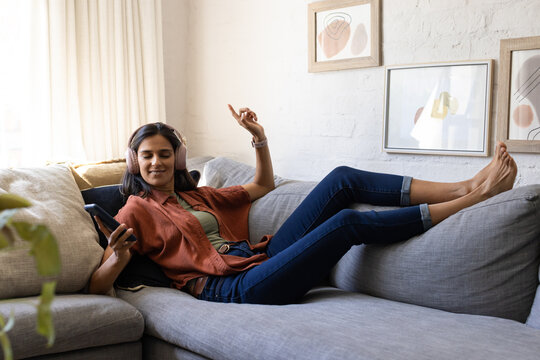 Adult woman reclining on gray sofa at home in rust shirt, jeans, holding phone, wearing headset