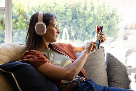 Indian woman in casual clothes reclining on sofa by window, using smartphone and wearing headphones