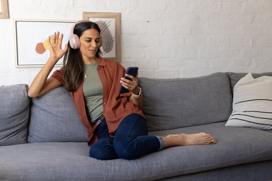 Indian woman sitting barefoot on grey sofa at home, wearing pink headphones and holding smartphone