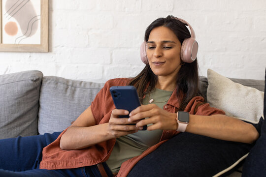 Woman reclining on gray sofa at home, wearing rust shirt and pink headphones, holding smartphone