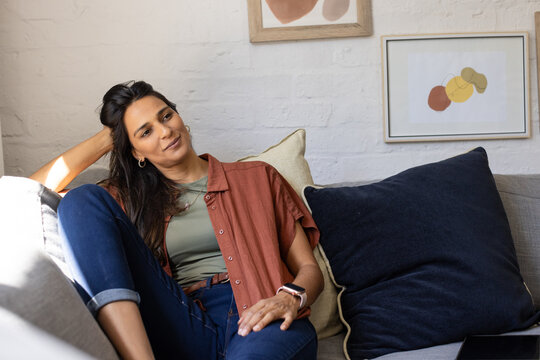 Woman reclining on gray sofa in living room, wearing rust shirt and smartwatch, checking tablet
