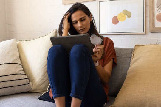 Indian woman sitting on sofa at home, holding tablet and credit card, wearing rust top