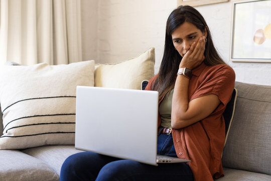 Adult Indian woman sitting on gray sofa working on silver laptop in home, copy space