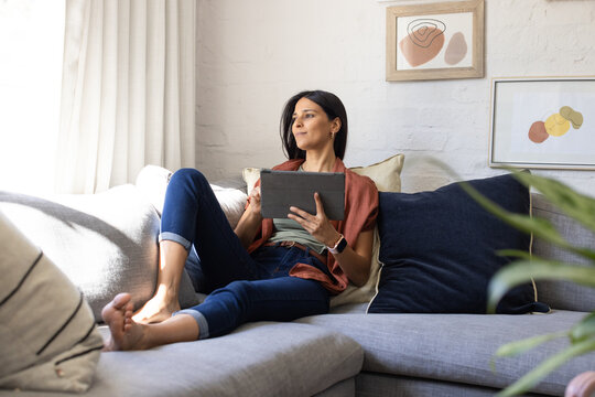 Adult Indian woman reclining on sectional sofa at home holding tablet, wearing jeans and smartwatch
