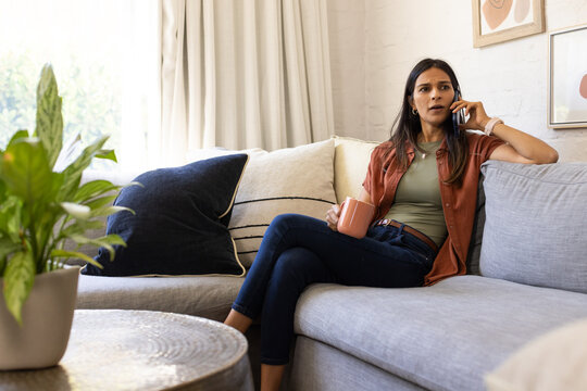 Indian woman in rust jacket sitting right on sofa holding mug, talking on smartphone at home