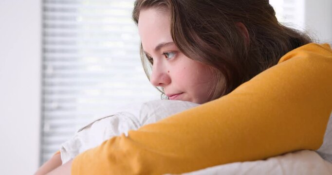 Woman in mustard-top at home looking past blinds, resting chin on pillow, seeking calm, copy space